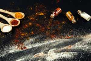 A variety of colorful spices, Salt and Vinegar Seasoning and measuring spoons are arranged on a black surface.